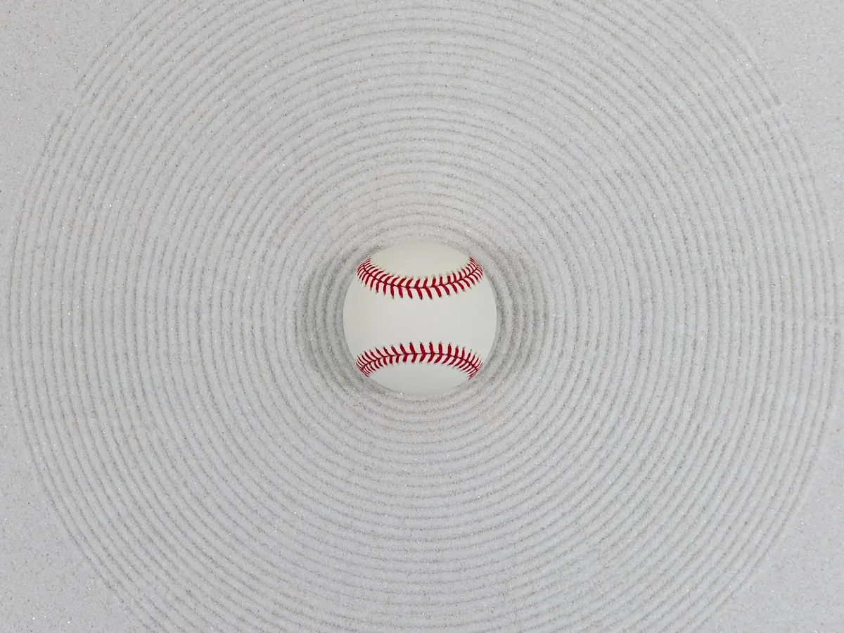 A plain white baseball with red stitching resting in raked sand